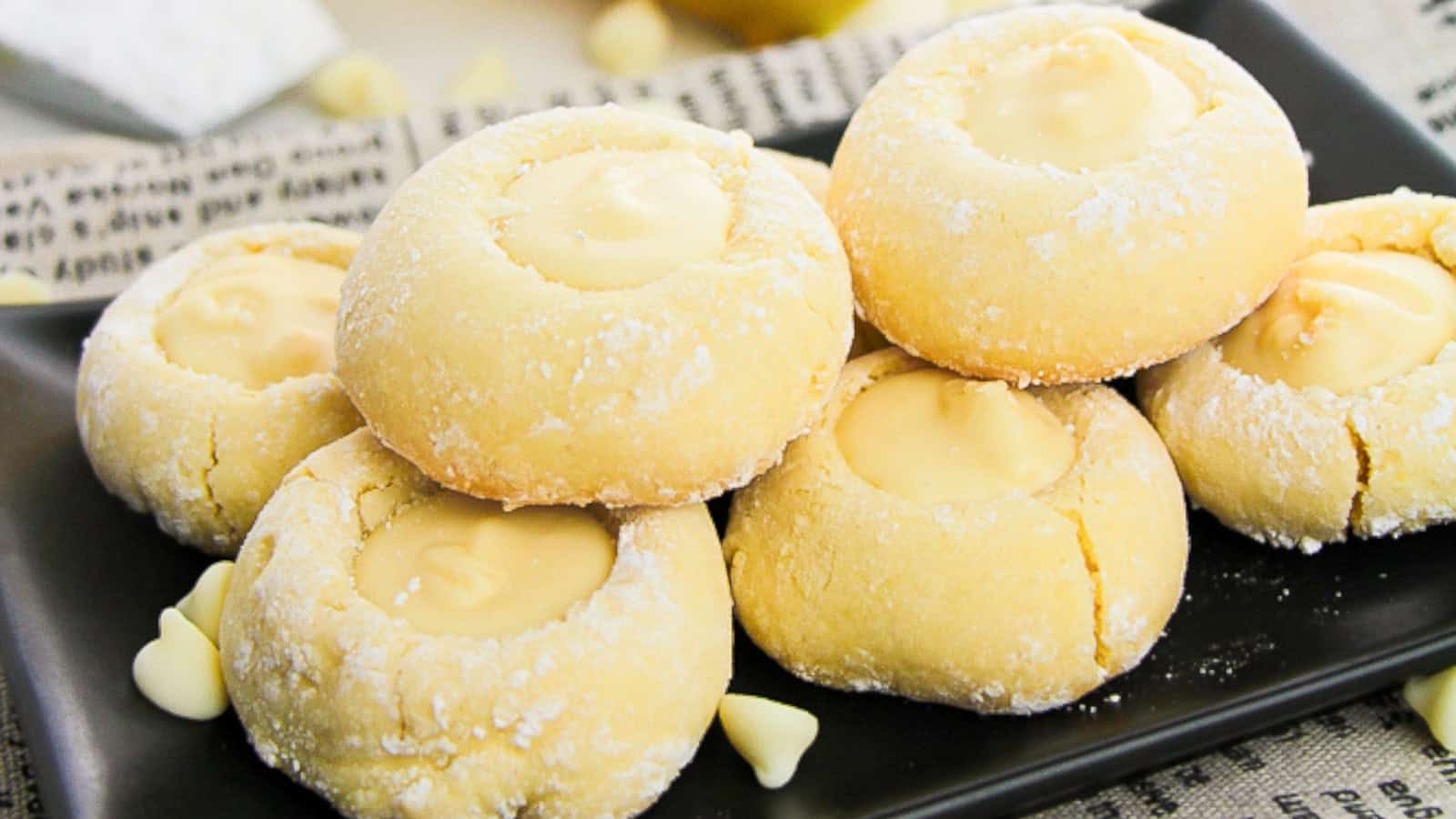 A plate of round, yellow thumbprint cookies dusted with powdered sugar, each filled with creamy white chocolate.