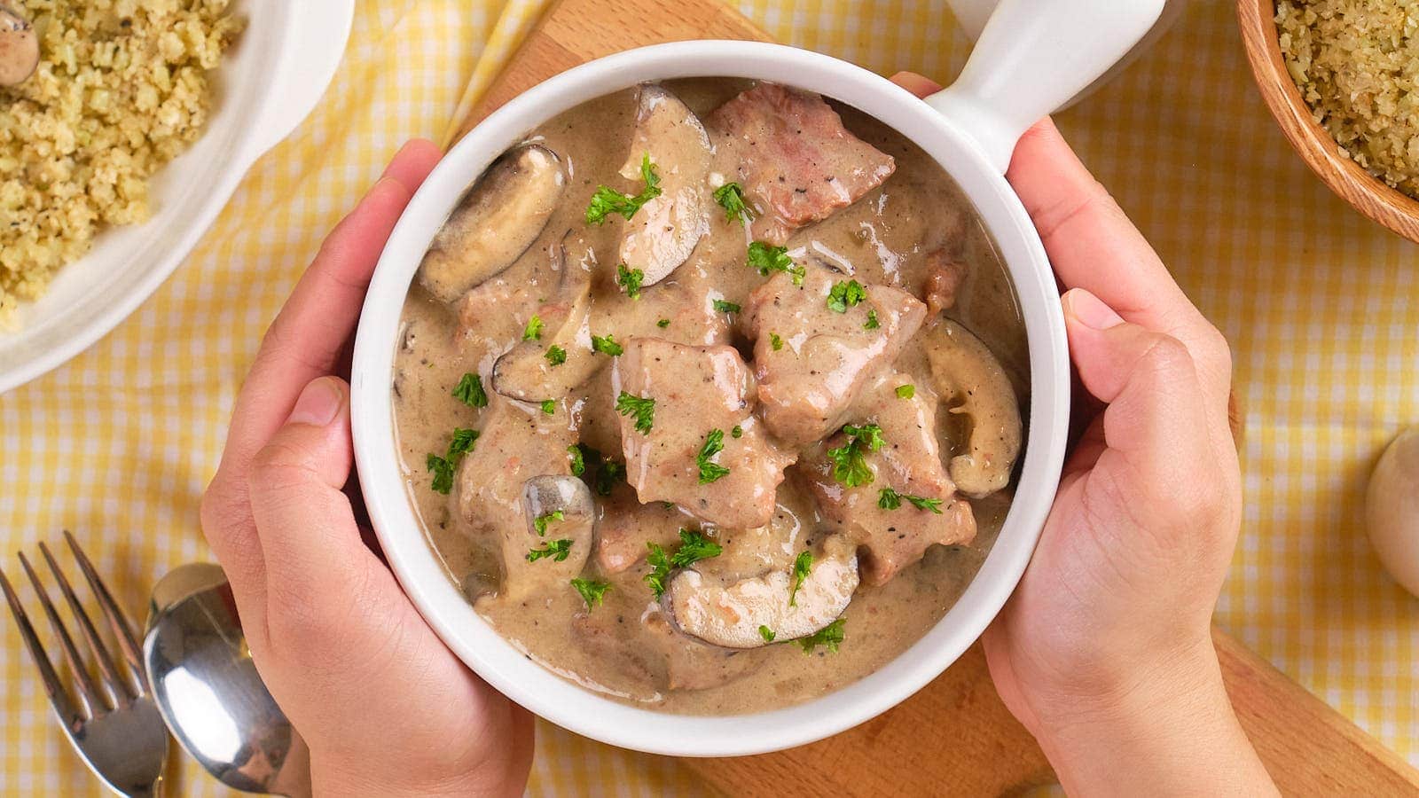 A person holds a bowl of beef stroganoff with mushrooms and creamy sauce, garnished with chopped parsley, on a checkered tablecloth.