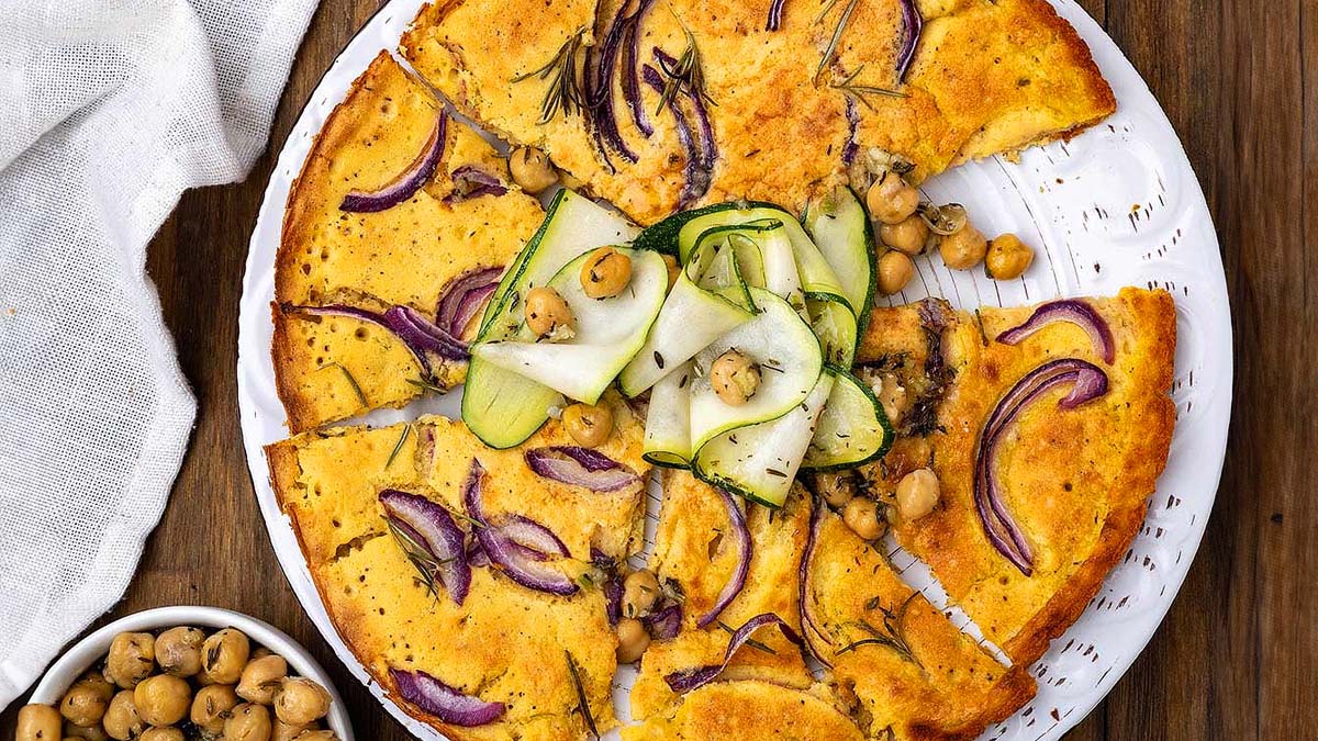 Chickpea flatbread topped with red onion, fresh rosemary, chickpeas, and zucchini ribbons, served on a white plate with a small bowl of chickpeas beside it.