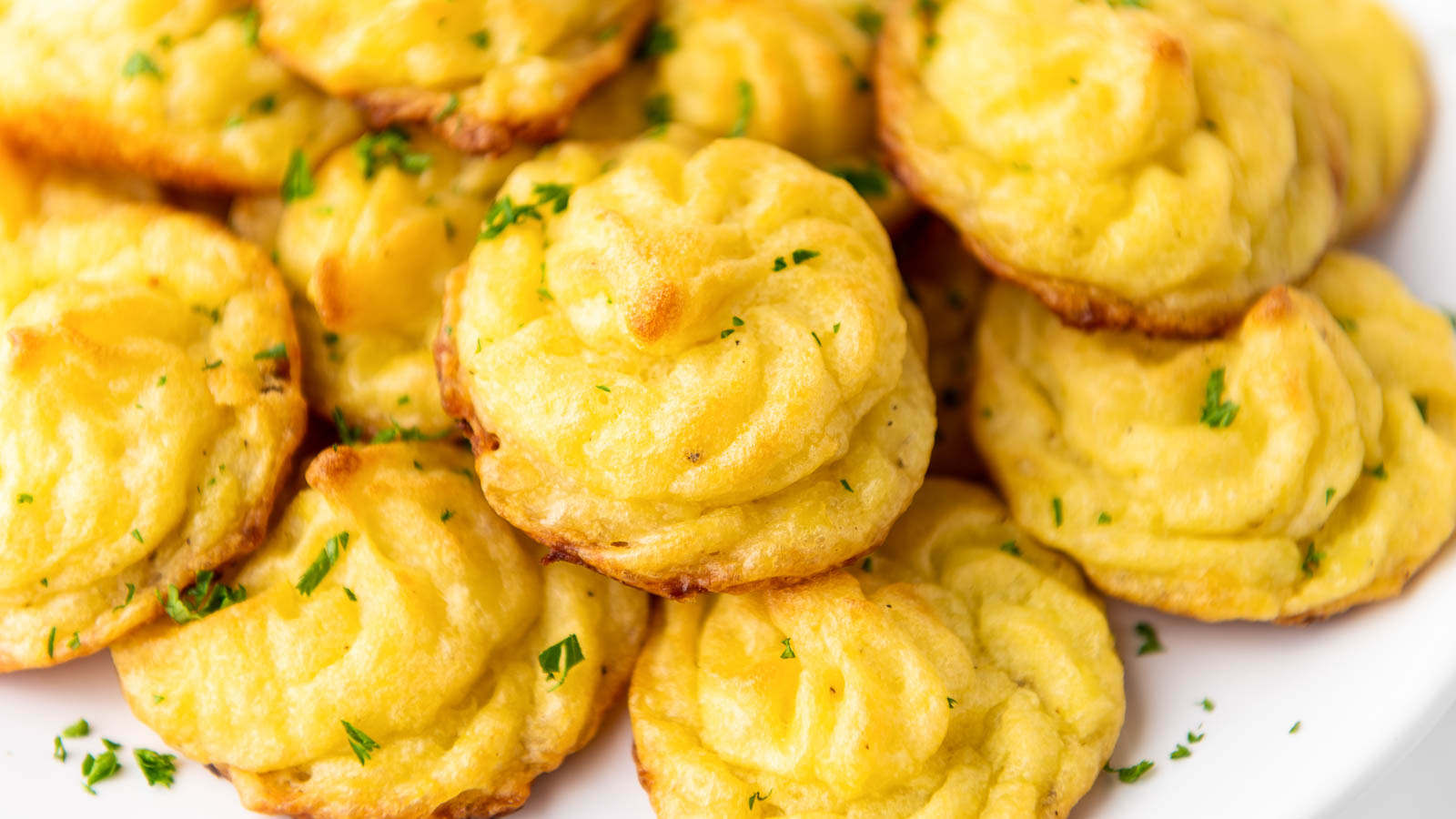 A close-up of several golden-brown duchess potatoes garnished with chopped parsley on a white plate.