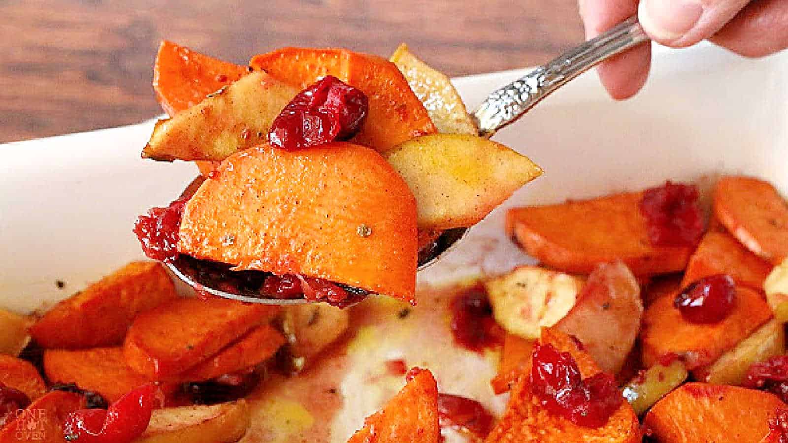 A spoon scoops baked sweet potatoes, apples, and cranberries from a white dish.