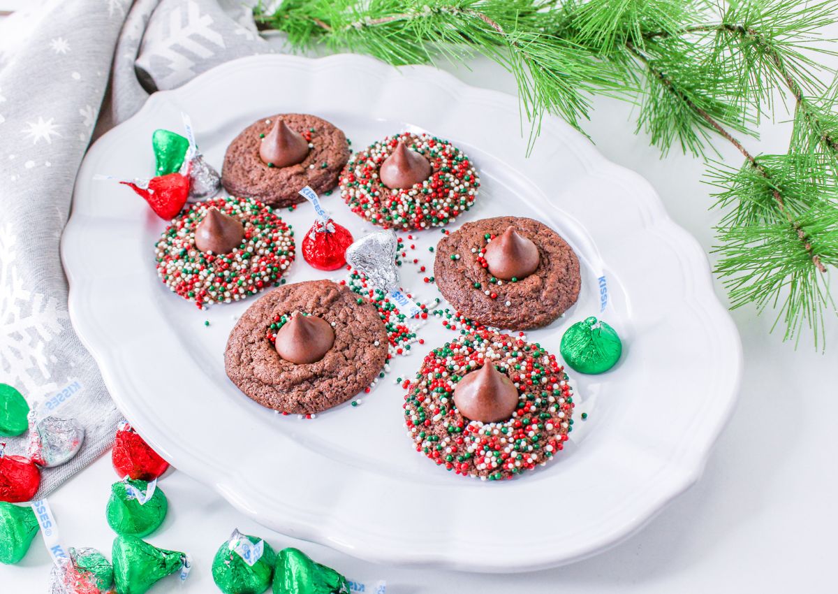 A white plate holds six chocolate cookies, some with Hersheyโs Kisses and festive sprinkles, surrounded by assorted Hersheyโs Kisses and pine branches.