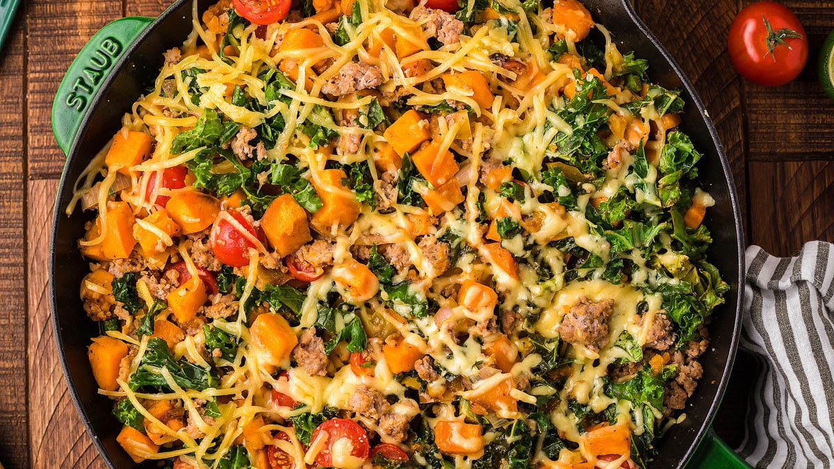 A skillet filled with a baked dish containing ground meat, kale, diced sweet potatoes, cherry tomatoes, and melted shredded cheese.