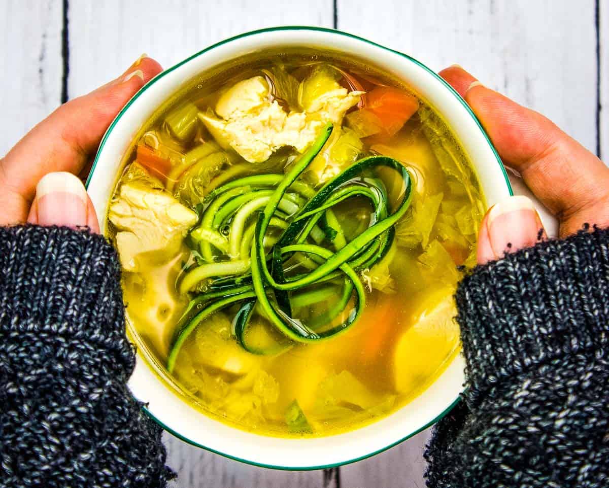 A person holding a bowl of chicken soup with zucchini noodles, carrots, and other vegetables, on a white wooden surface.