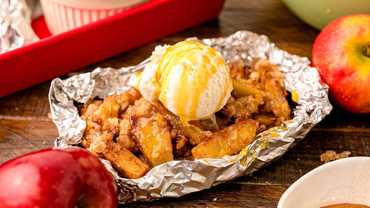 Baked apple slices with crumble topping in foil, topped with a scoop of vanilla ice cream and caramel drizzle, surrounded by fresh apples.