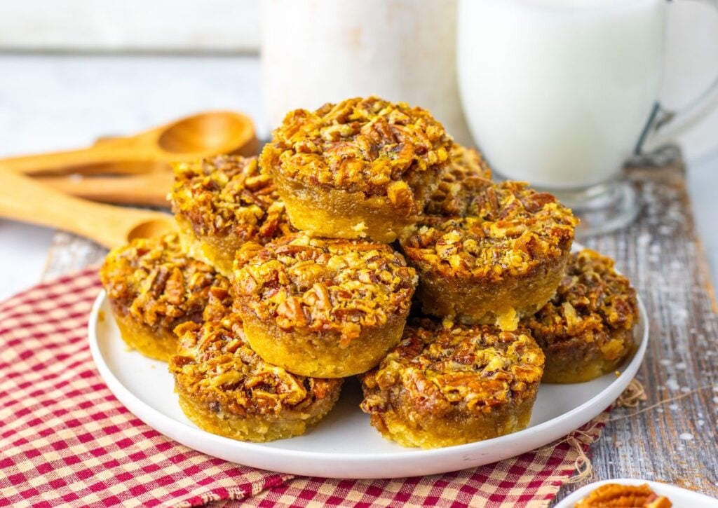 A plate of pecan pie muffins is stacked on a white dish, with a glass of milk and wooden measuring spoons in the background on a red-checkered cloth.