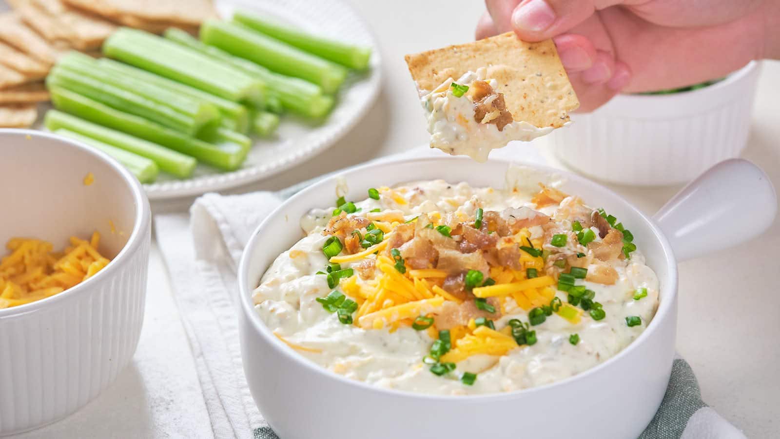 A hand dips a cracker into a bowl of creamy dip topped with bacon, shredded cheese, and chives; celery sticks and crackers are on a plate nearby.