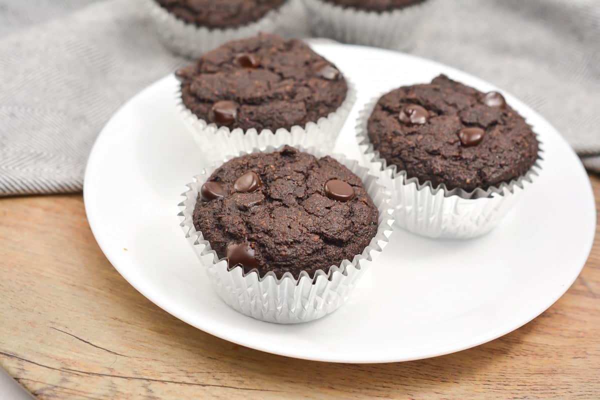 Three chocolate chip muffins in white paper liners are arranged on a white plate, with a gray cloth and more muffins in the background.