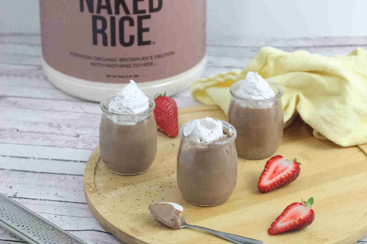 Three small glass jars of chocolate mousse topped with whipped cream are on a wooden board with sliced strawberries, a spoon, and a container labeled “Naked Rice” in the background.