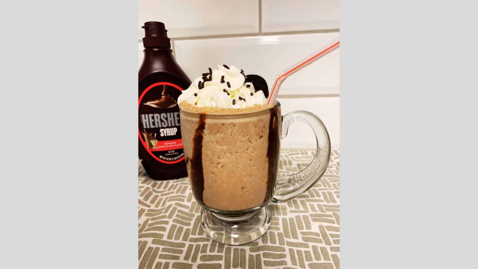 A glass mug of chocolate milkshake topped with whipped cream, chocolate sprinkles, and a cookie, with a bottle of Hershey’s syrup in the background.