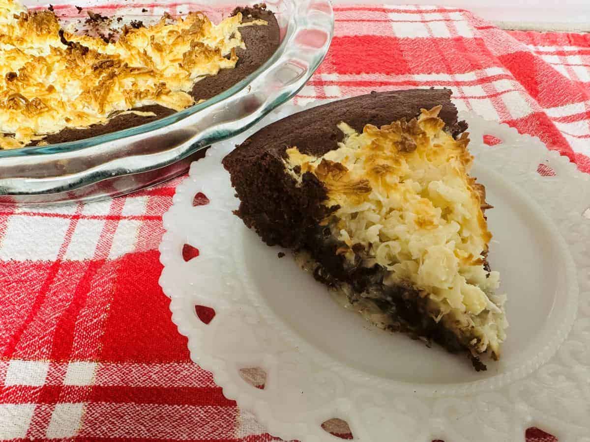 A slice of chocolate cake with a toasted coconut topping on a white plate, next to the remaining cake in a glass pie dish, on a red and white checkered tablecloth.