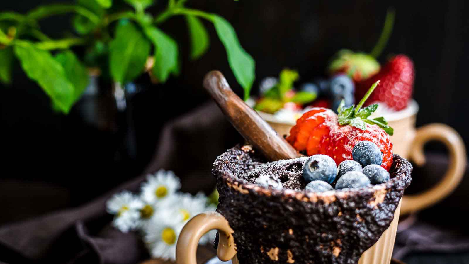 A chocolate mug cake topped with blueberries, a strawberry, and a chocolate stick, with another dessert and flowers blurred in the background.