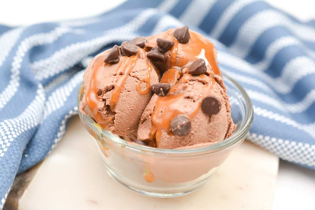 A glass bowl with three scoops of chocolate ice cream topped with chocolate chips and caramel sauce, set in front of a blue and white striped cloth.