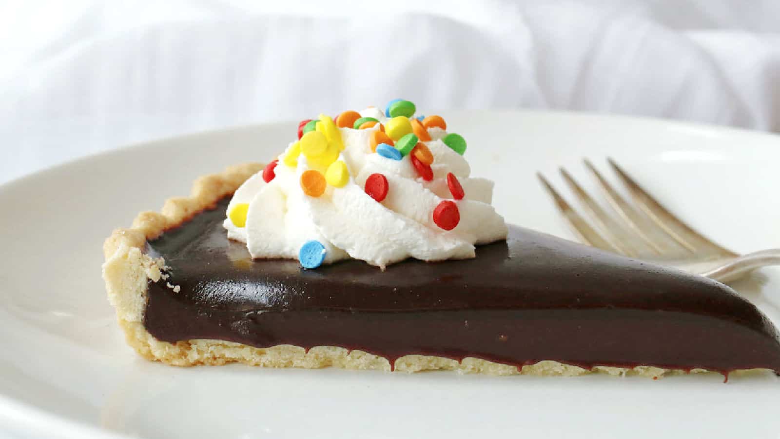A slice of chocolate pie topped with whipped cream and colorful round sprinkles sits on a white plate with a fork.