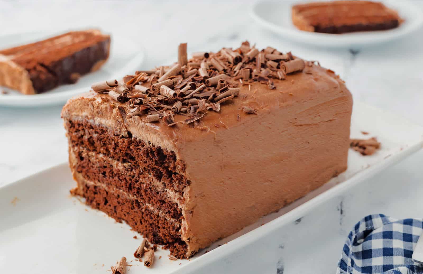 A rectangular chocolate layer cake with chocolate frosting and chocolate shavings on top, served on a white platter with two slices in the background.