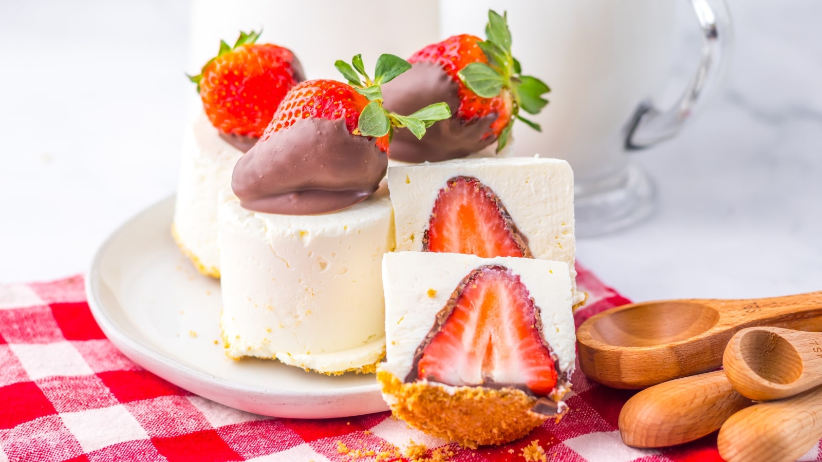 Mini cheesecakes topped with chocolate-covered strawberries on a white plate, with a red checkered cloth and wooden spoons nearby.