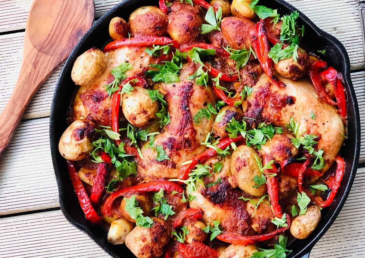 A skillet filled with roasted chicken, baby potatoes, red bell pepper strips, and fresh parsley, placed on a wooden surface beside a wooden spoon.
