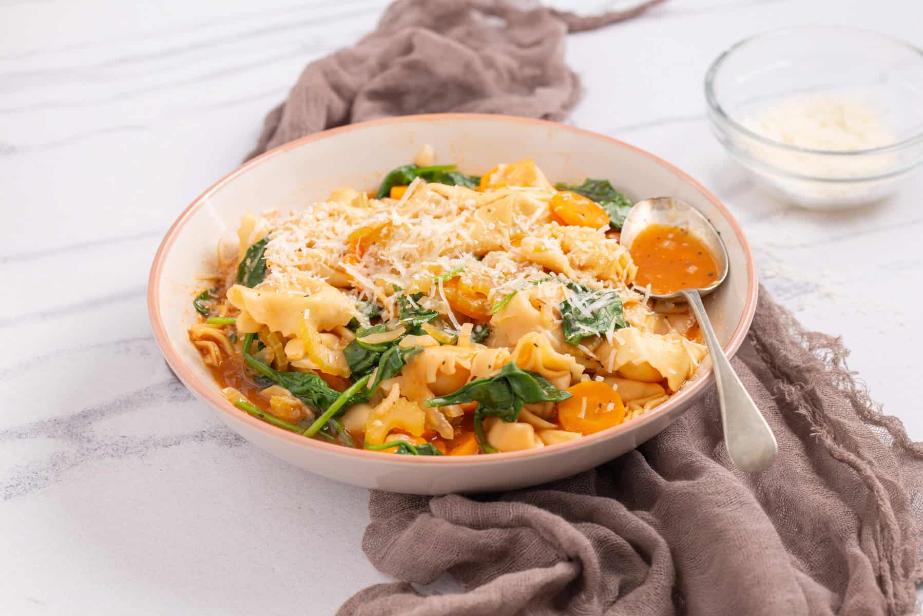 A bowl of pasta with yellow tomatoes, spinach, sauce, and grated cheese on top, with a spoon and a small bowl of extra cheese nearby.