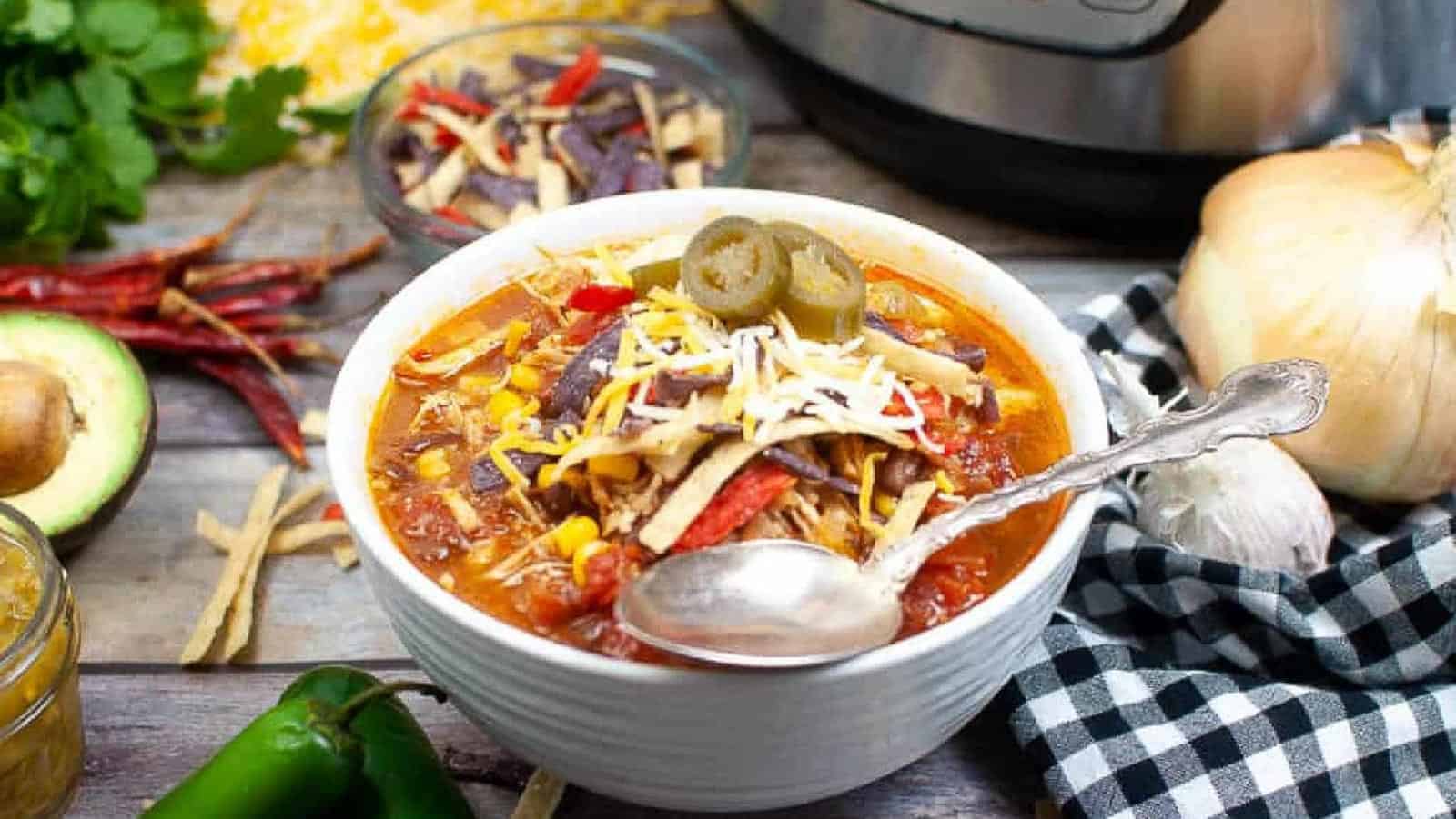 A bowl of chicken tortilla soup topped with shredded cheese, tortilla strips, and jalapeños, with a spoon in the bowl and various fresh ingredients around it.