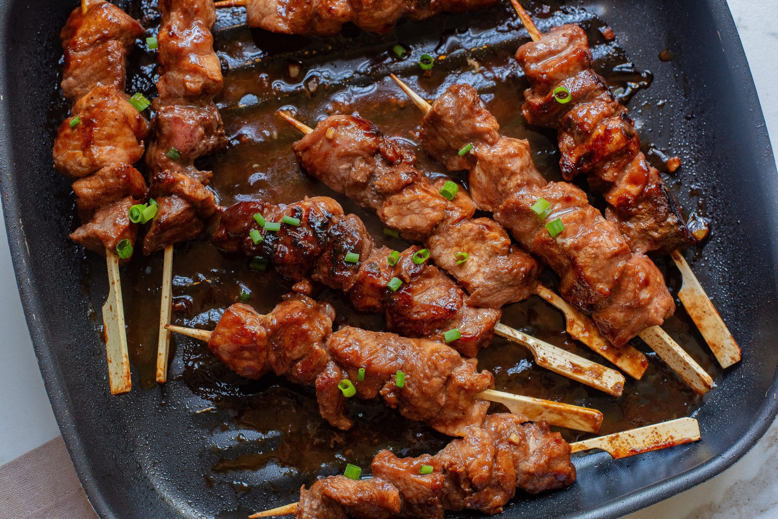 Grilled meat skewers with sauce and chopped green onions on a black pan.