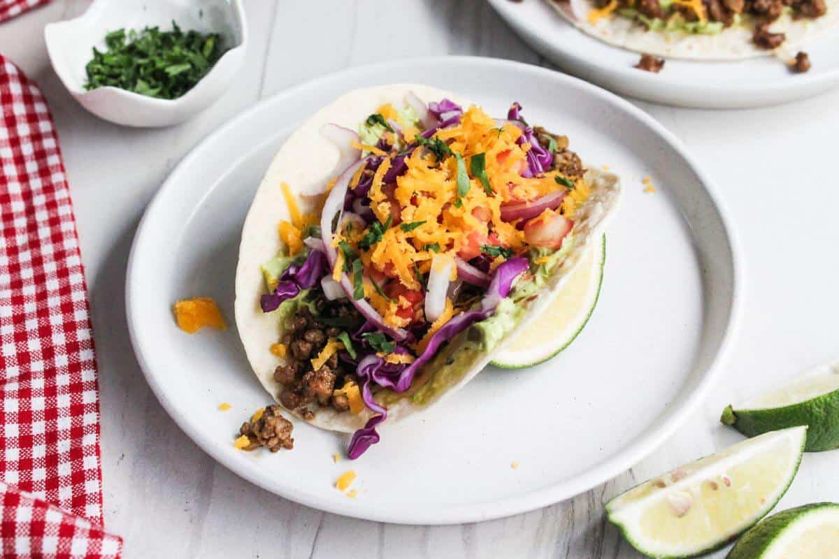 A taco on a white plate is filled with ground meat, shredded cheese, cabbage, onions, and tomato, served with a lime wedge.