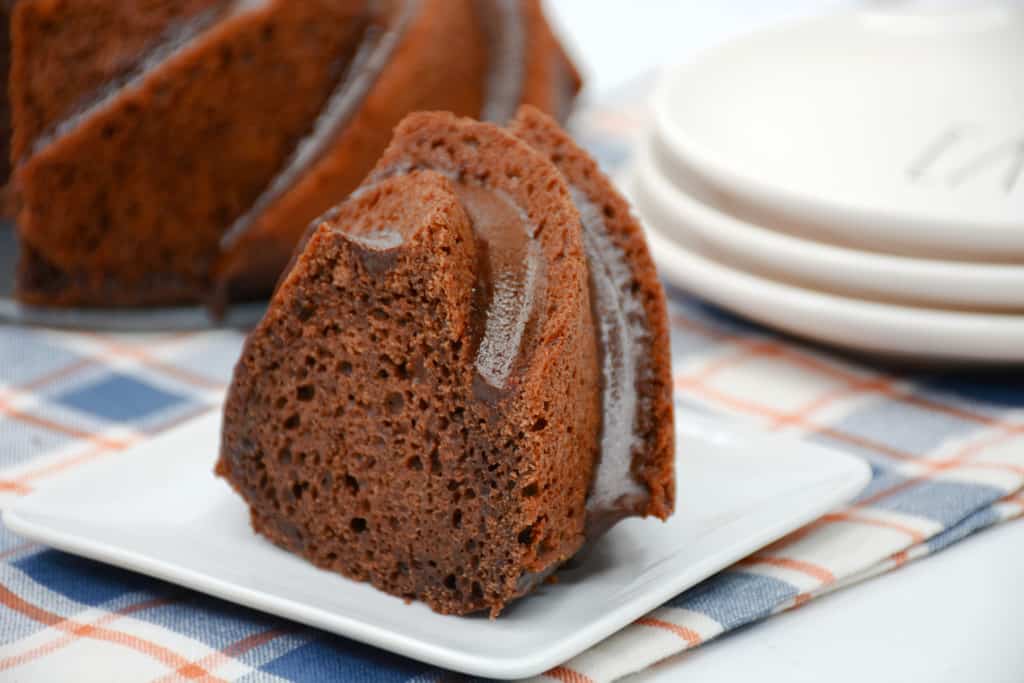 A slice of chocolate bundt cake with chocolate glaze on a square white plate, set on a plaid cloth with stacked plates in the background.