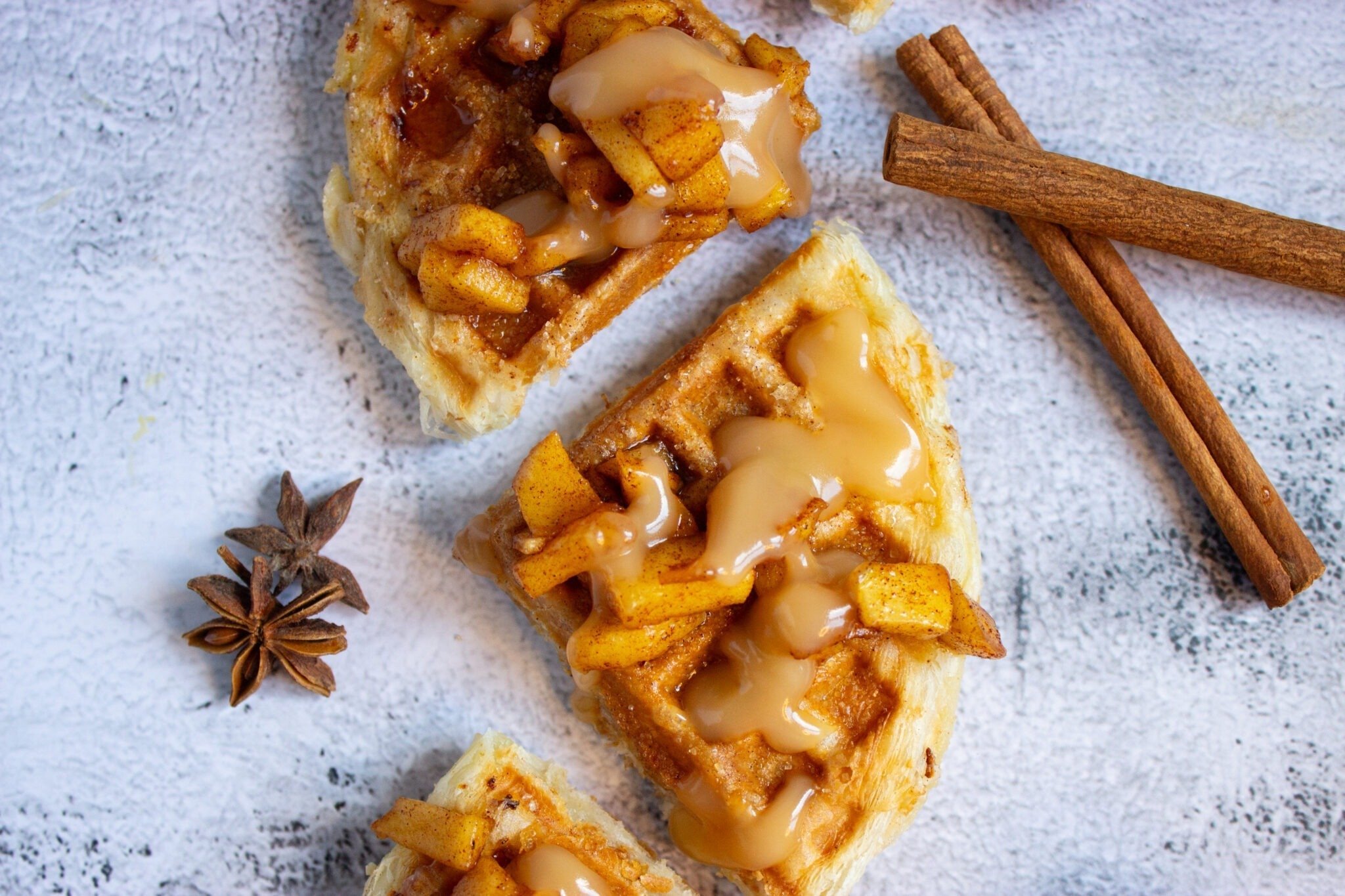 Waffle pieces topped with spiced apple chunks and sauce are arranged on a surface, next to whole cinnamon sticks and a star anise.