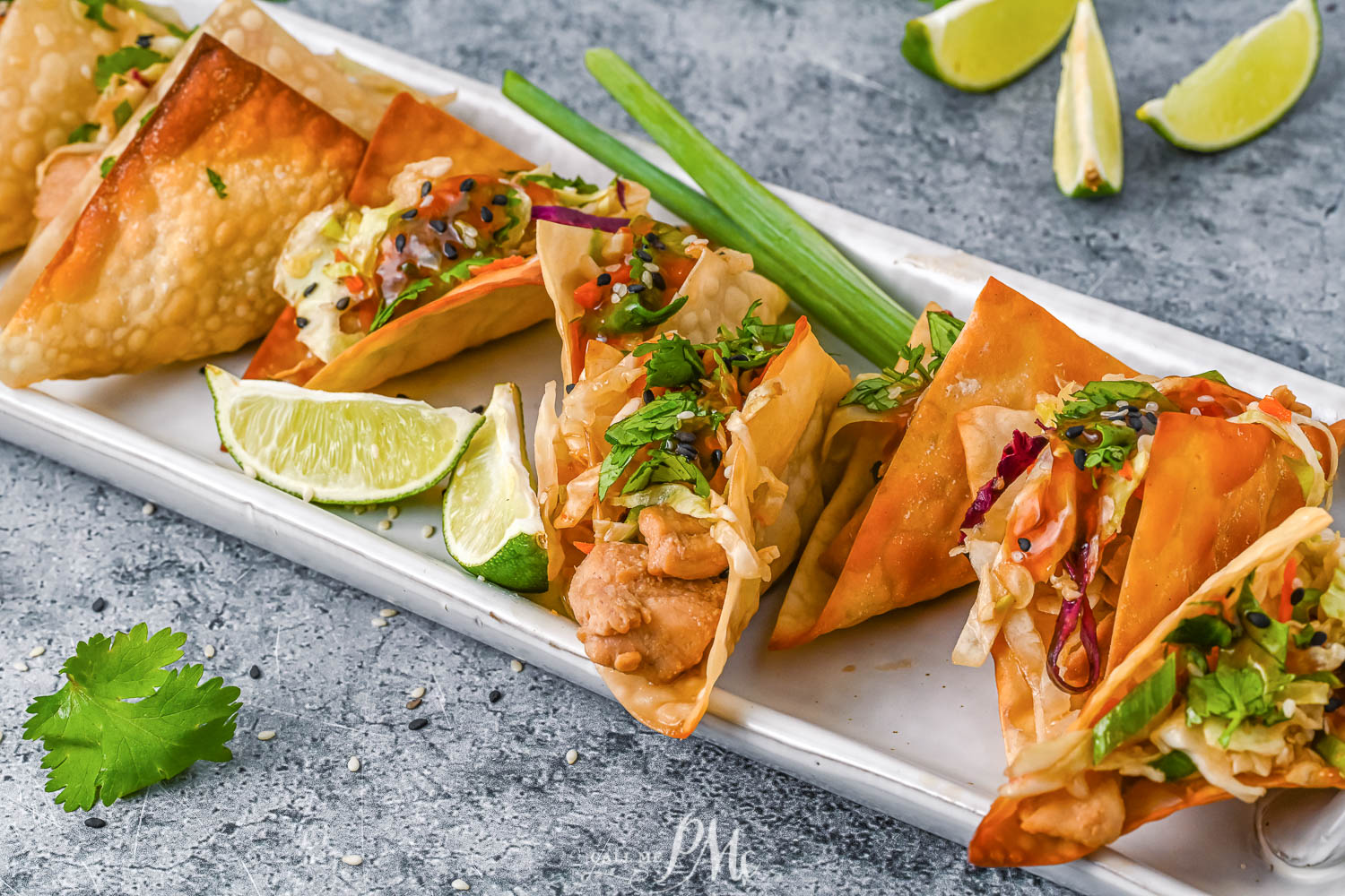 A rectangular plate with crispy wonton tacos filled with chicken, vegetables, and garnished with cilantro and sesame seeds, served with lime wedges.