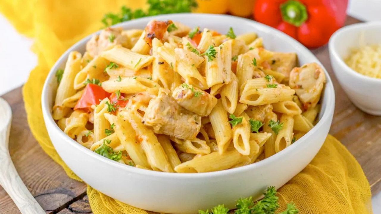 A white bowl filled with penne pasta, grilled chicken pieces, red bell peppers, and fresh parsley on a wooden table.