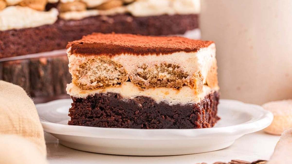 A slice of layered dessert with a brownie base, creamy filling, ladyfinger layer, and a dusting of cocoa powder on top, served on a white plate.
