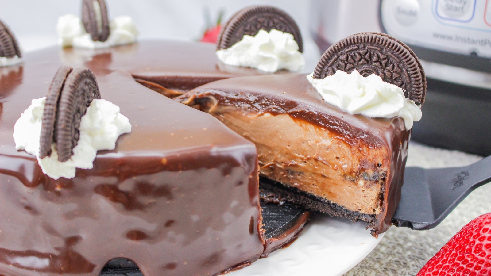 A chocolate cheesecake with an Oreo cookie crust, topped with chocolate glaze, whipped cream, and Oreo halves. A slice is being lifted from the cake.