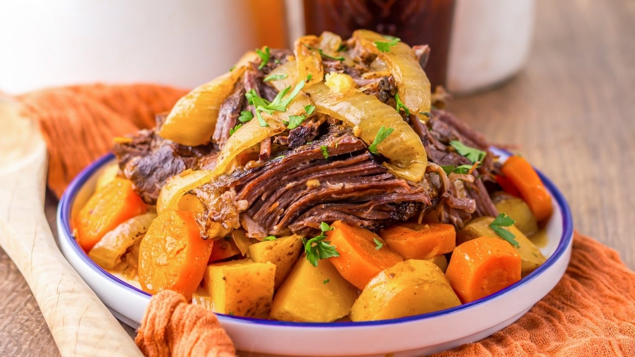 A plate of pot roast topped with onions, served with carrots and potatoes, garnished with chopped parsley.