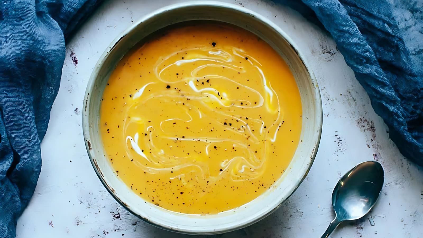 A bowl of creamy orange soup with a swirl of cream and a sprinkle of black pepper, placed next to a spoon and blue fabric on a light surface.
