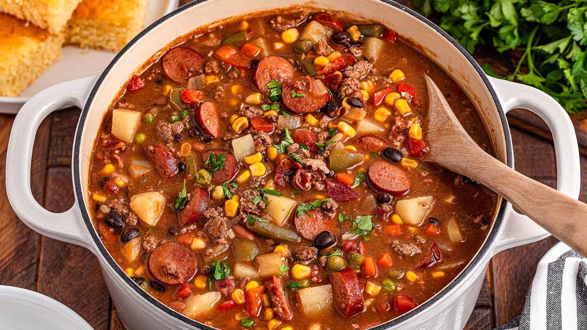 A pot of hearty stew with sausage, ground meat, corn, beans, potatoes, tomatoes, and vegetables, with a wooden spoon in the pot and cornbread on the side.