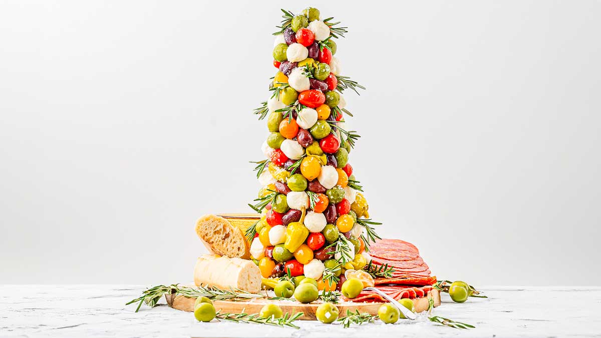 A Christmas tree-shaped arrangement of tomatoes, olives, and mozzarella balls on a wooden board, surrounded by sliced bread, cured meats, and loose olives.