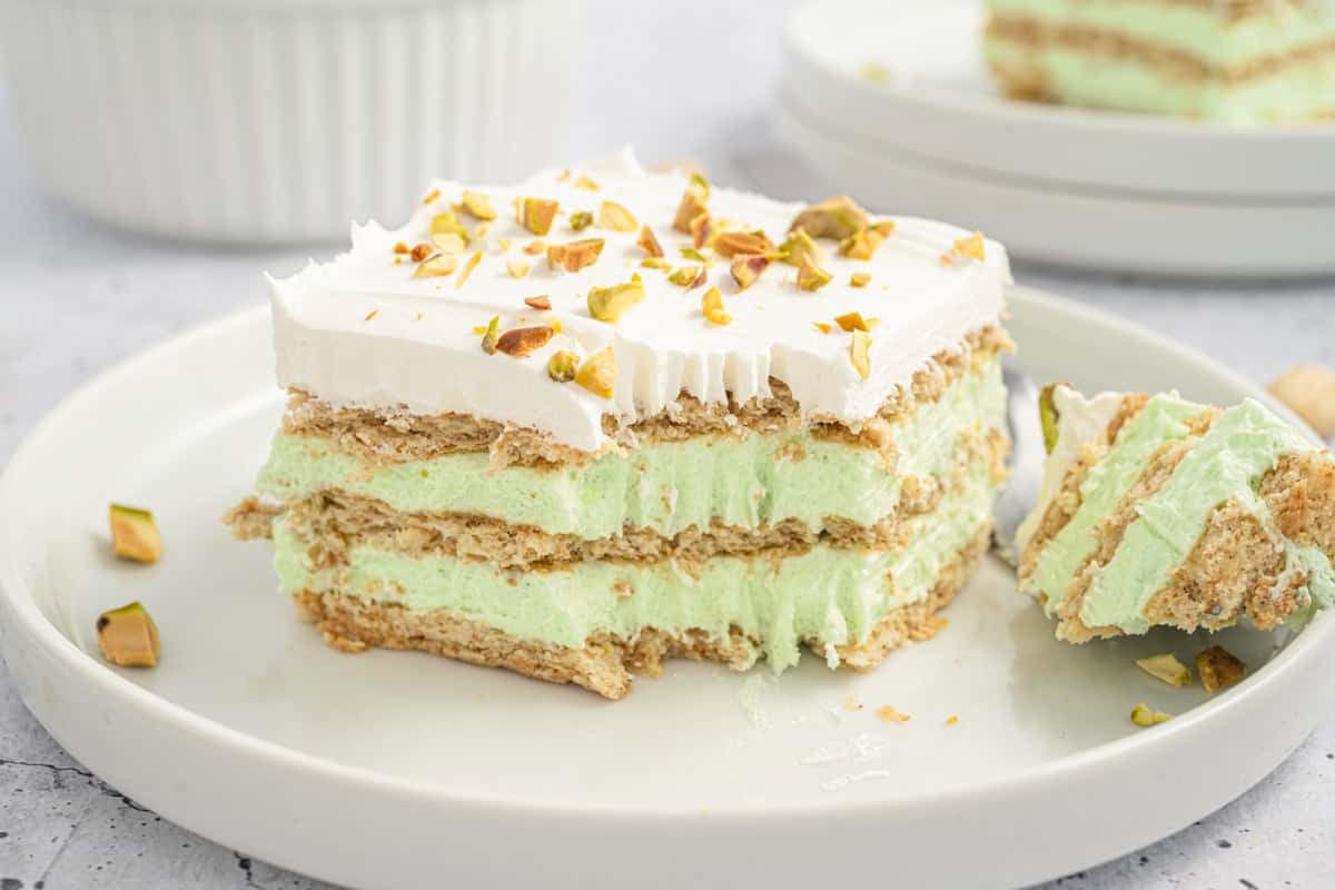 A slice of layered dessert with green filling, whipped topping, crushed pistachios, and a partially eaten piece on a white plate.