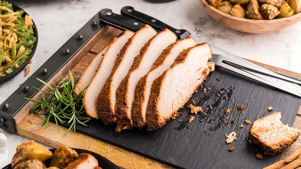 Sliced roasted turkey breast arranged on a slate serving board with a sprig of rosemary, surrounded by side dishes.