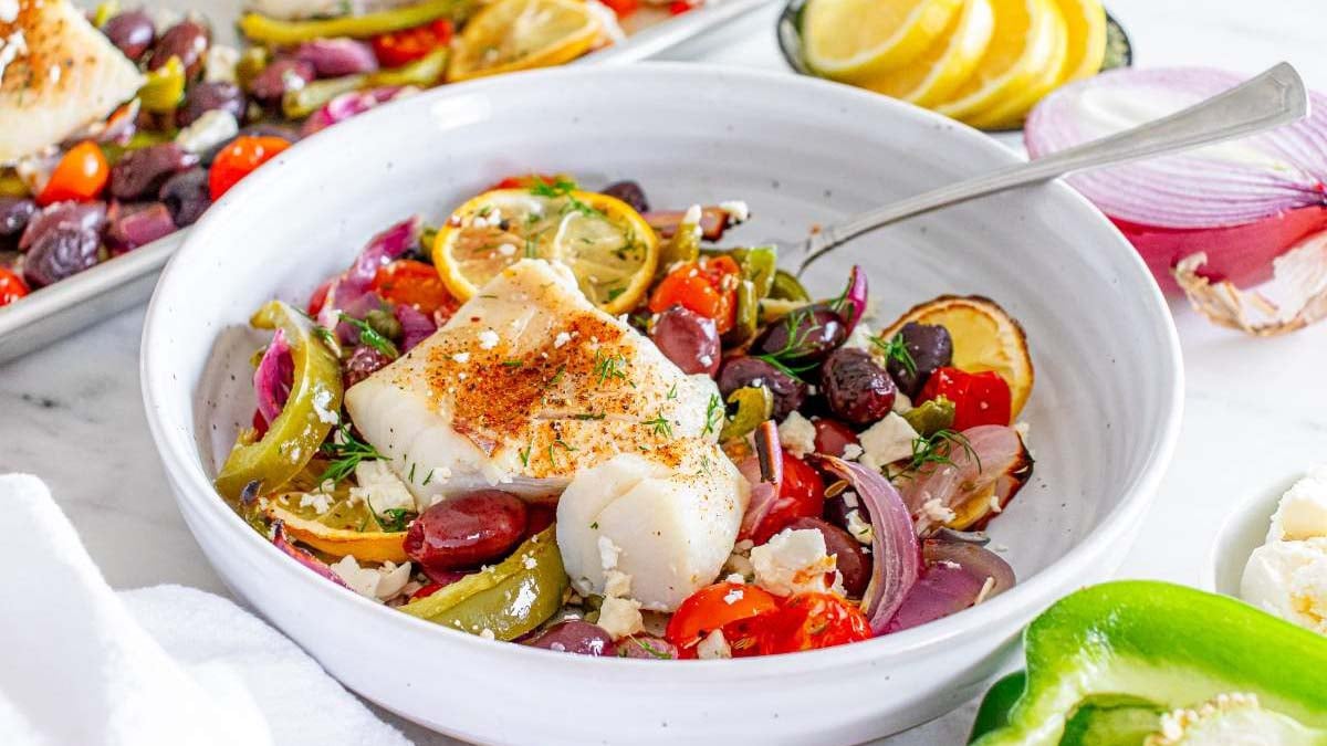A white bowl with baked fish, lemon slices, cherry tomatoes, olives, red onion, green pepper, and herbs, with a tray of similar food and lemon slices in the background.