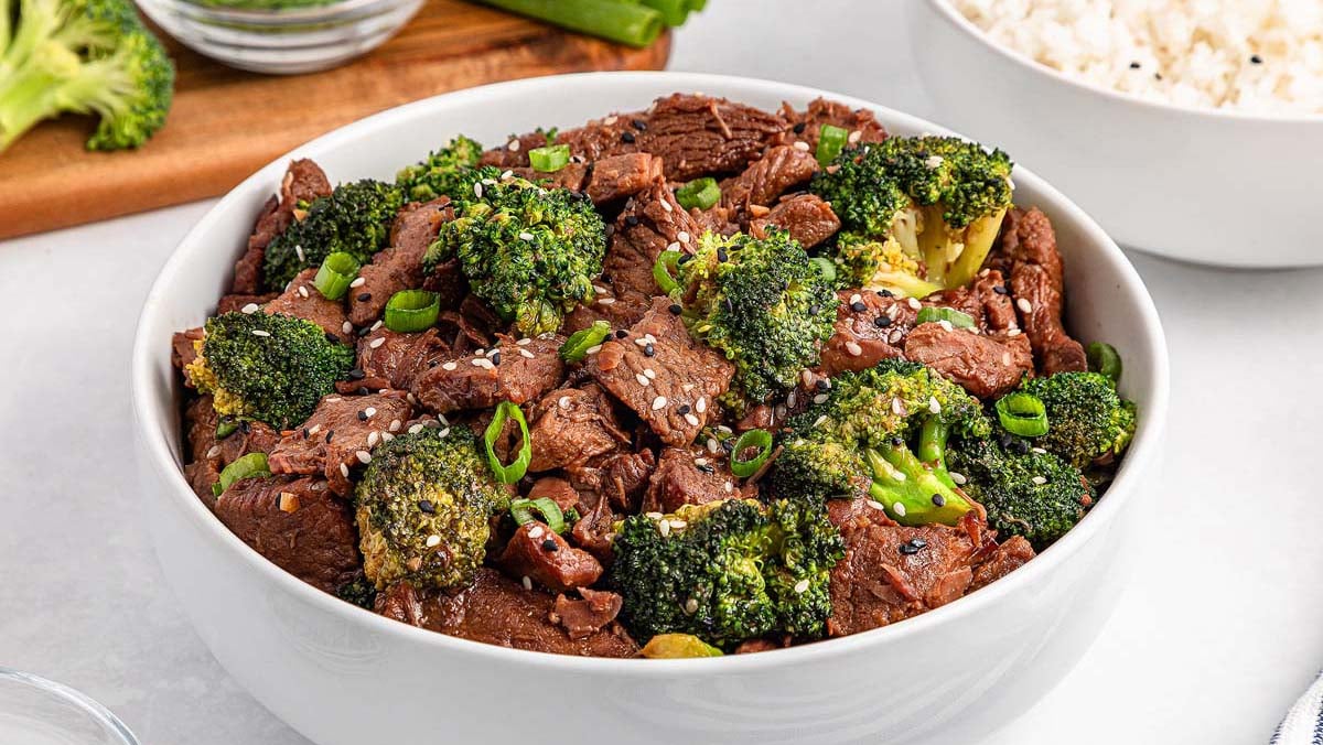 A white bowl filled with beef and broccoli stir-fry garnished with sesame seeds and green onions; rice and vegetables are visible in the background.