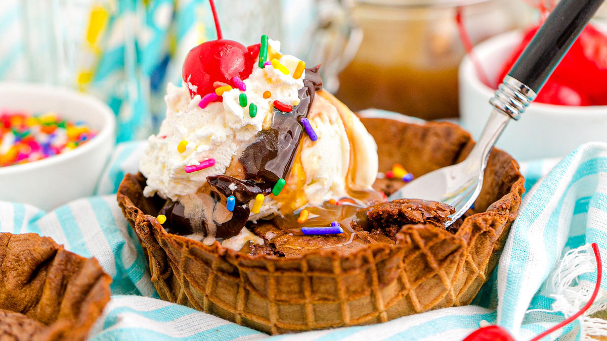 A waffle bowl filled with brownie, vanilla ice cream, whipped cream, chocolate syrup, sprinkles, and a cherry, with a spoon on the side.