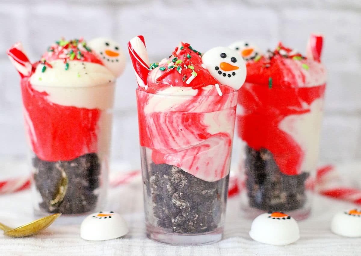 Three clear cups filled with crushed cookies, swirled red and white cream, festive sprinkles, candy canes, and snowman face decorations on top.