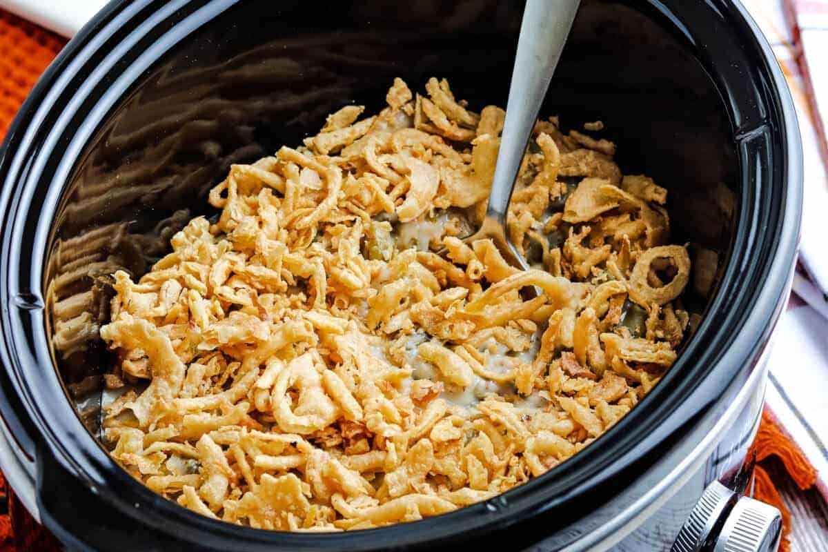 A slow cooker filled with green bean casserole topped with crispy fried onions, with a serving spoon inside.