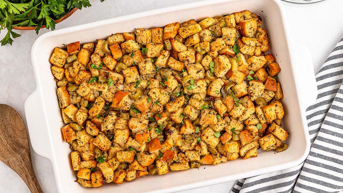 A white baking dish filled with golden-brown cubed bread stuffing garnished with herbs, resting on a white surface next to a striped towel and a wooden spoon.