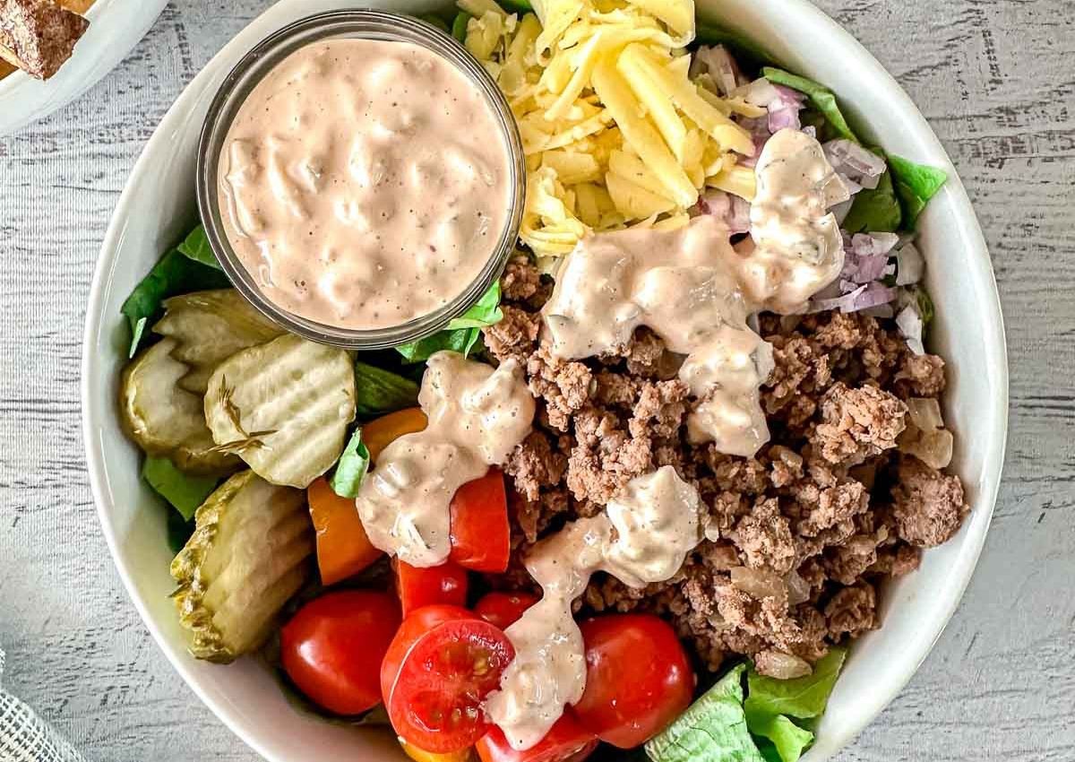 A bowl with ground beef, shredded cheese, chopped onions, sliced pickles, cherry tomatoes, lettuce, and a side of creamy dressing.
