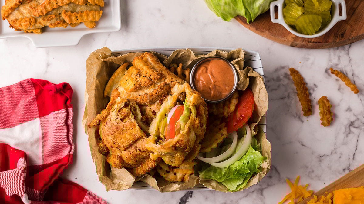 A basket with a fried chicken sandwich, crinkle-cut fries, lettuce, tomato, onion, and dipping sauce on parchment paper, with pickles and more fries on the side.