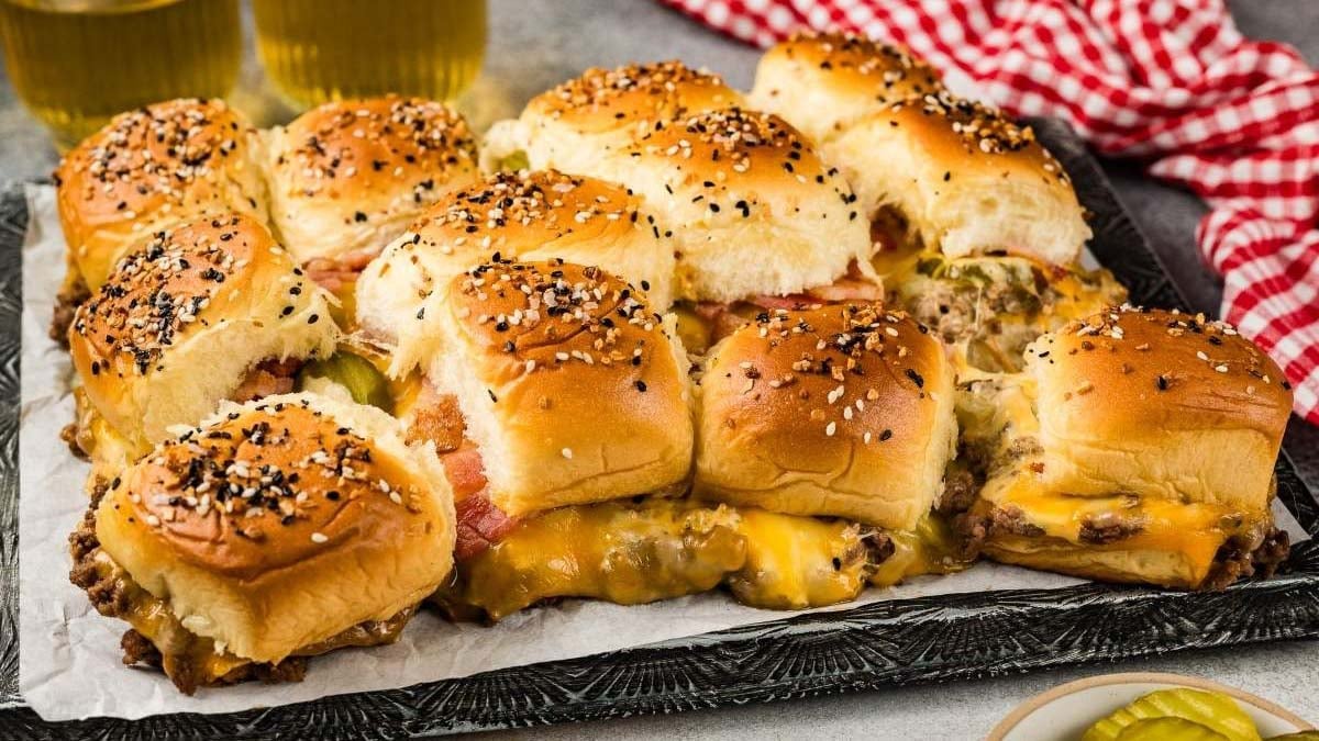A tray of baked cheeseburger sliders topped with sesame seeds, featuring melted cheese, pickles, and soft buns, with drinks and a red checkered cloth in the background.