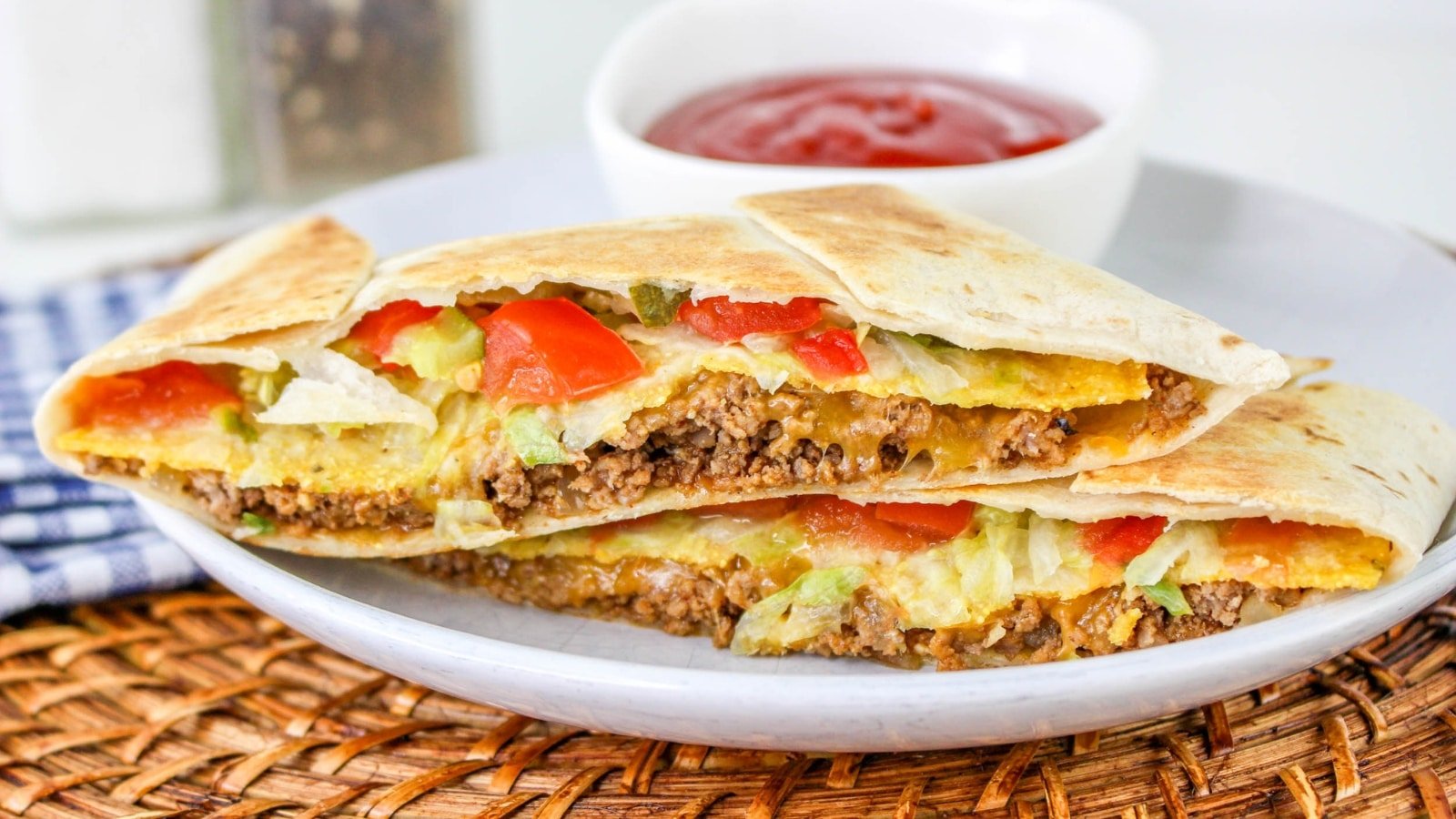A plate with two halves of a beef, cheese, lettuce, and tomato quesadilla, served with a small bowl of red dipping sauce.