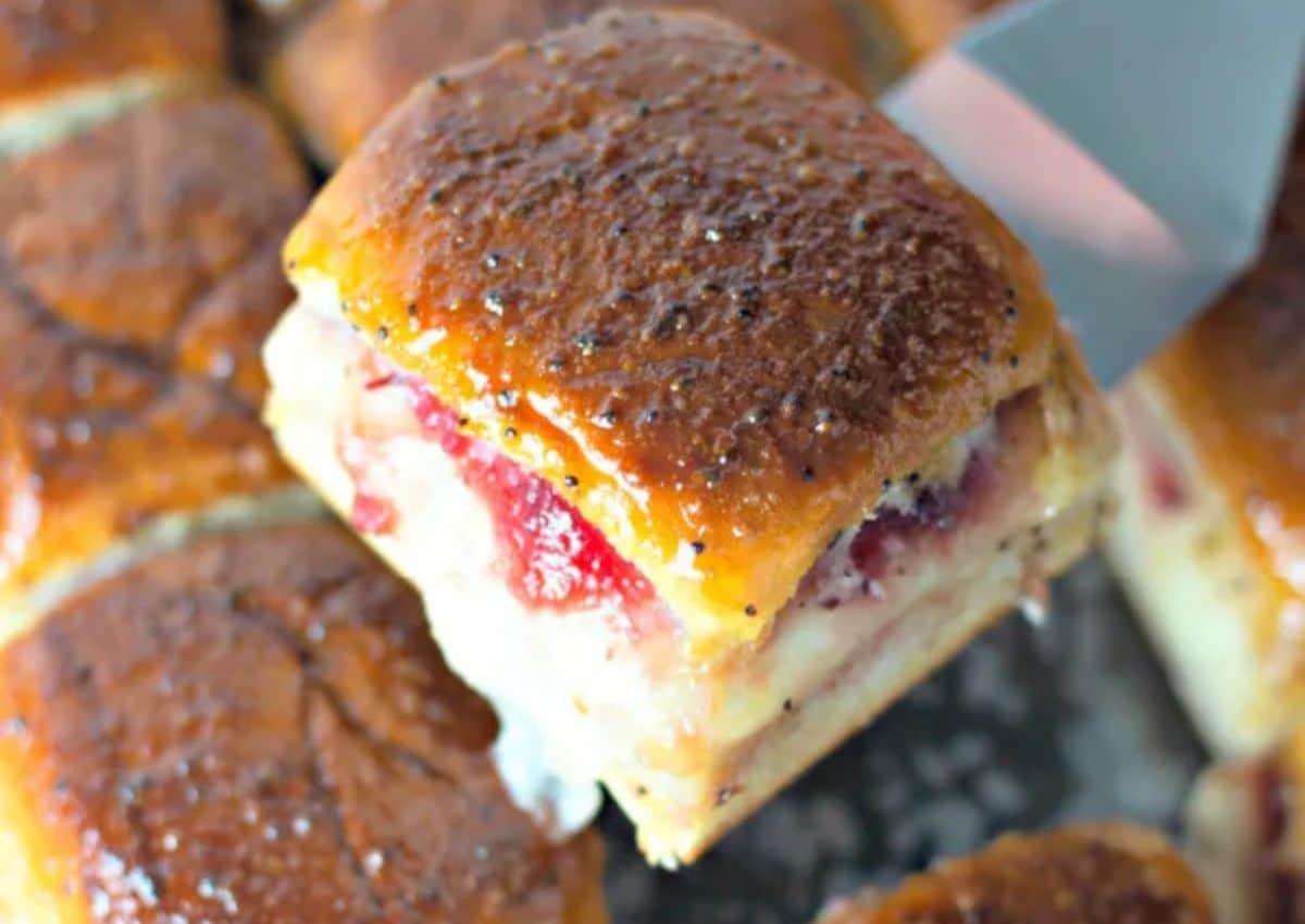 A close-up of a baked ham and cheese slider with a golden, glazed top being lifted from a tray, showing layers of melted cheese and ham.