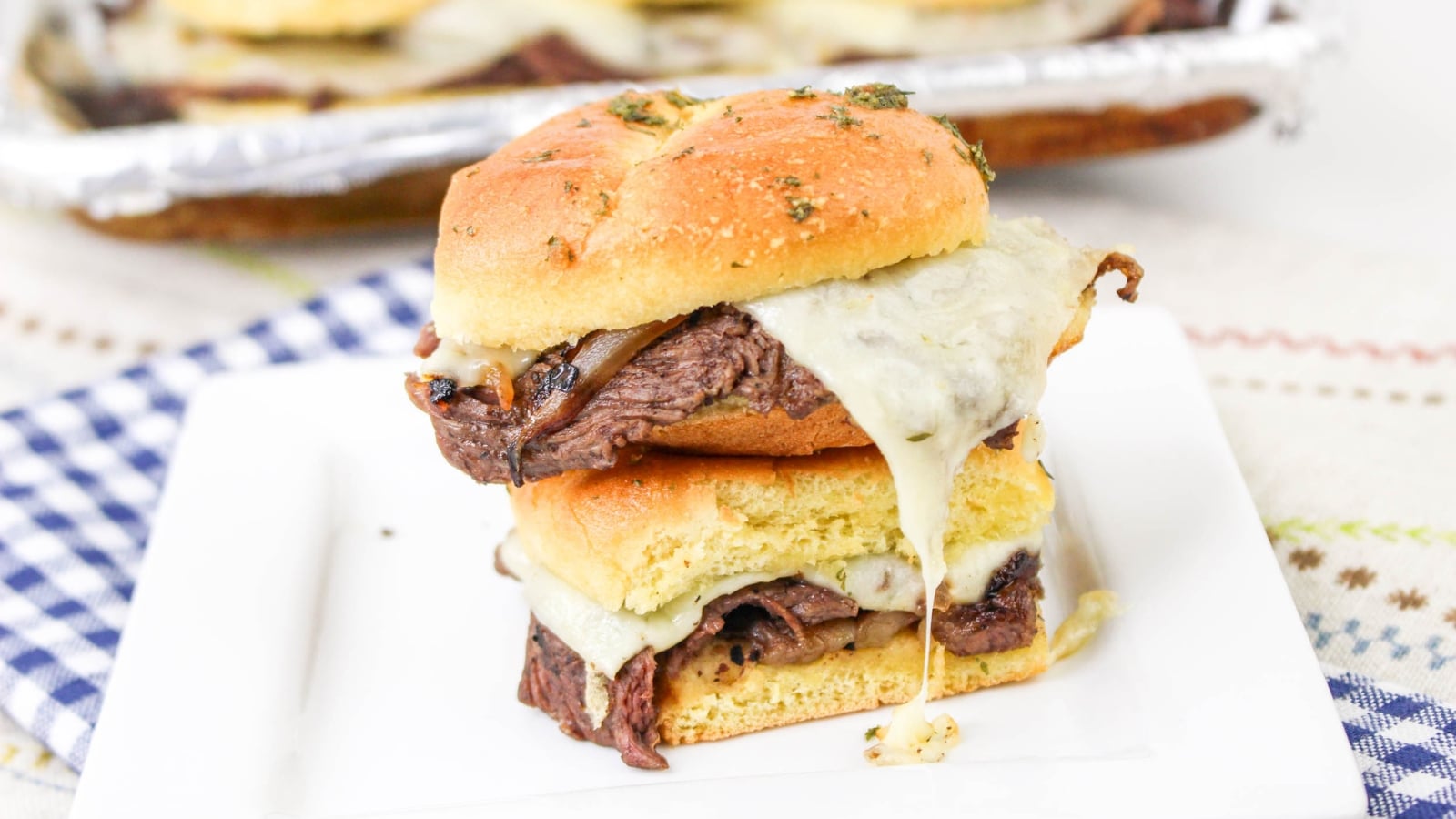 A steak sandwich with melted cheese and grilled onions on a toasted bun, served on a white plate.