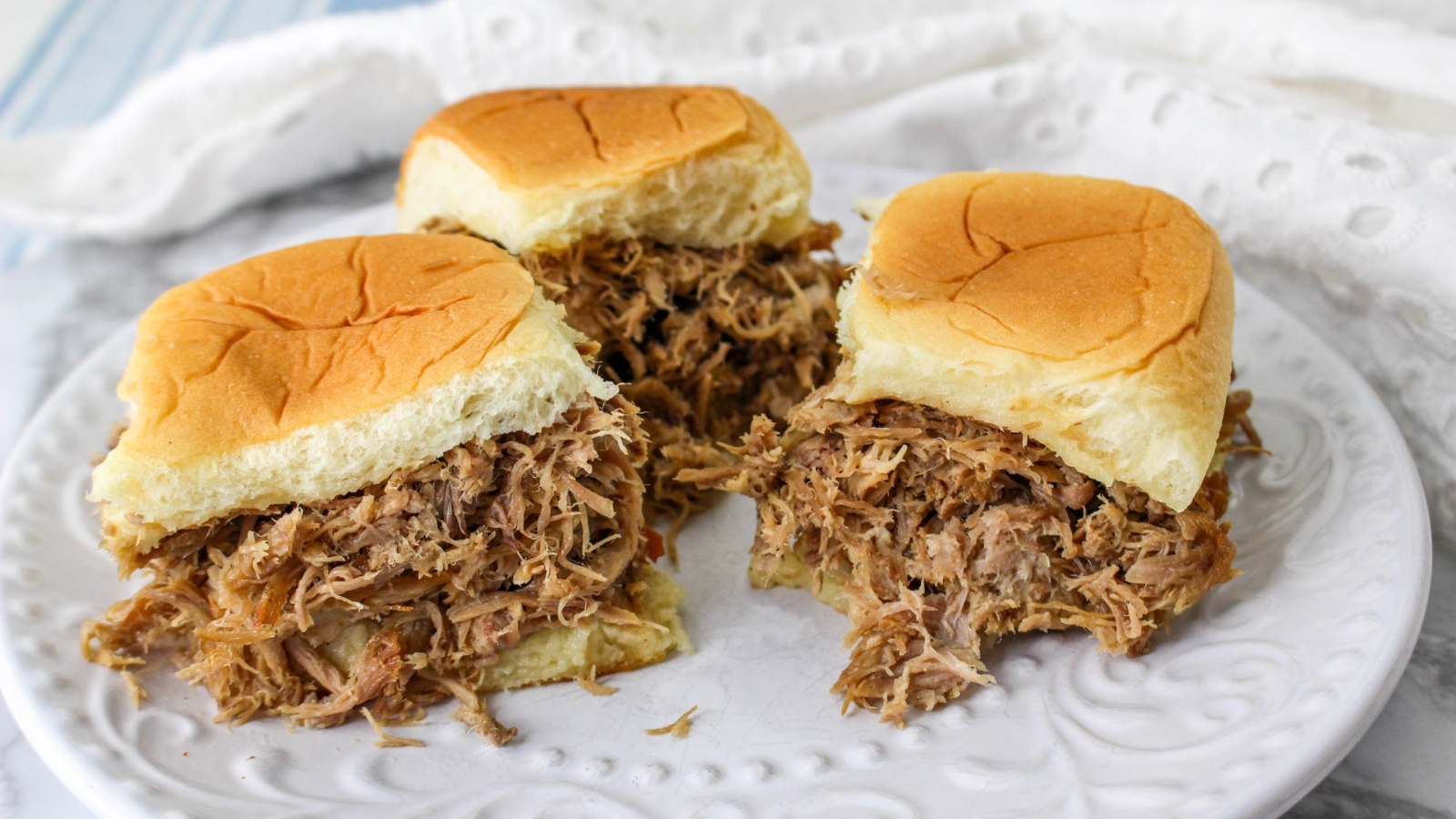 Three pulled pork sliders on a white plate, each with shredded pork filling and soft sandwich buns.